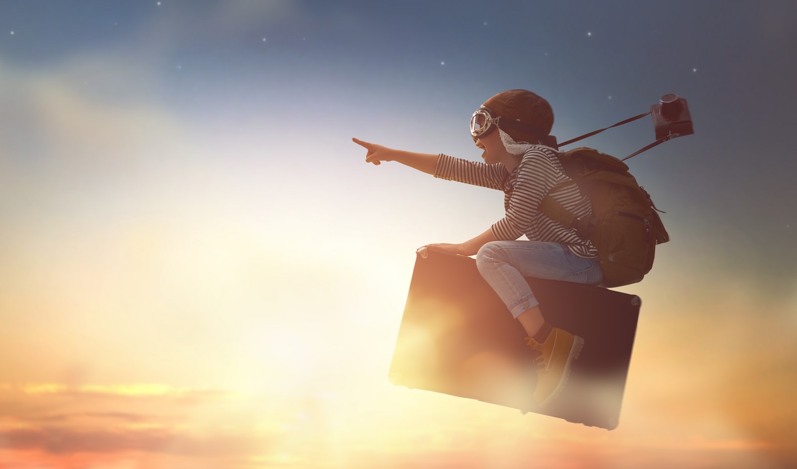 photo d’un enfant de profil avec un casque d’aviateur et un appareil photo sur une valise dans le ciel pointant le doigt droit devant lui